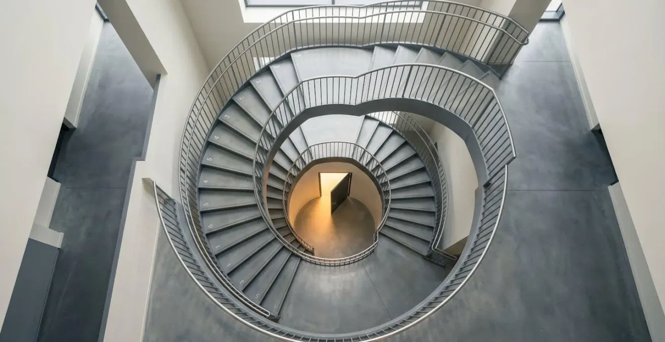 Vue aérienne d'un escalier en colimaçon avec marches numérotées et lumière naturelle descendante, porte dorée au bas symbolisant l'opportunité